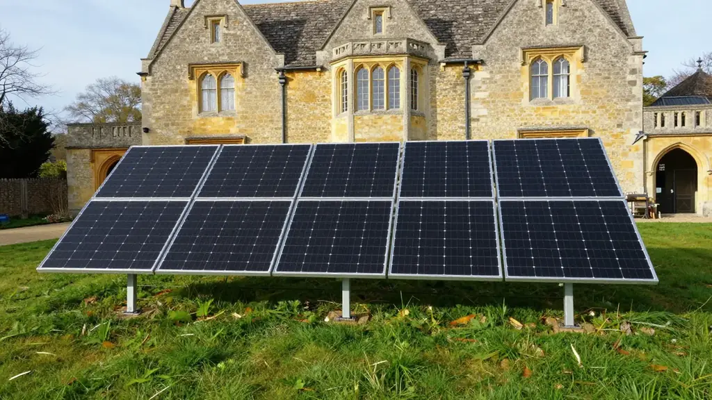 A ground-mounted array of black solar panels on a lawn with a historic stone manor house behind