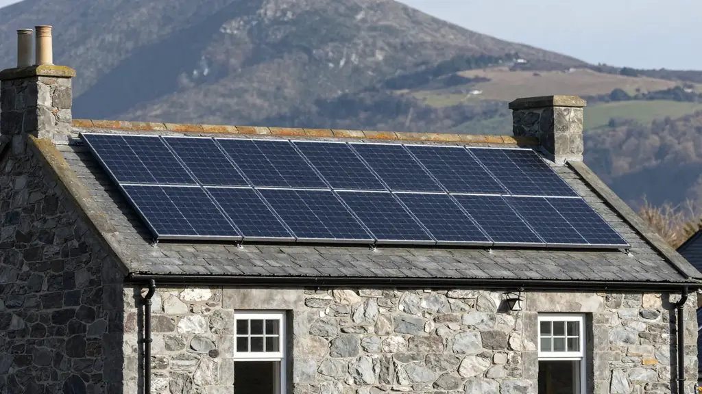 A low black solar array on the grey slate roof of a stone cottage with hills rising behind