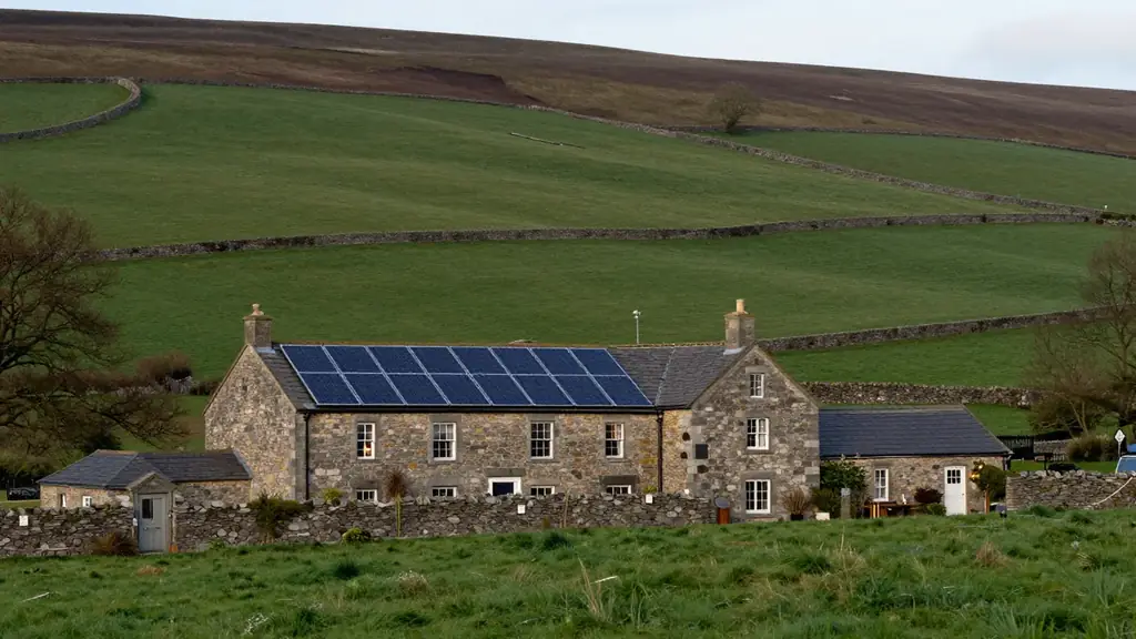 A stone farmhouse with a black solar array on its roof, set among green fields and hills